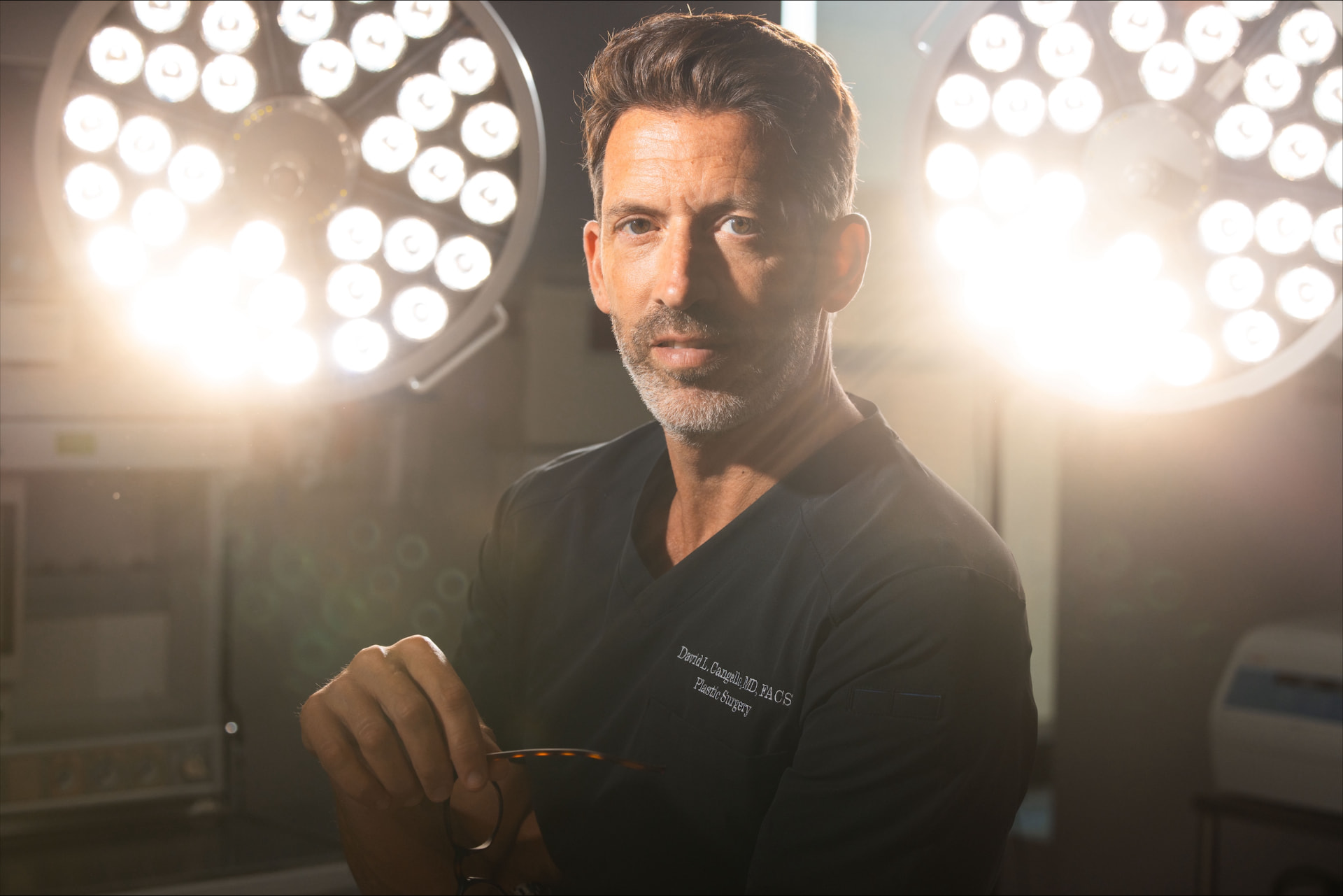 Dr. David Cangello in black surgical scrubs standing between illuminated operating room lights at his Manhattan practice